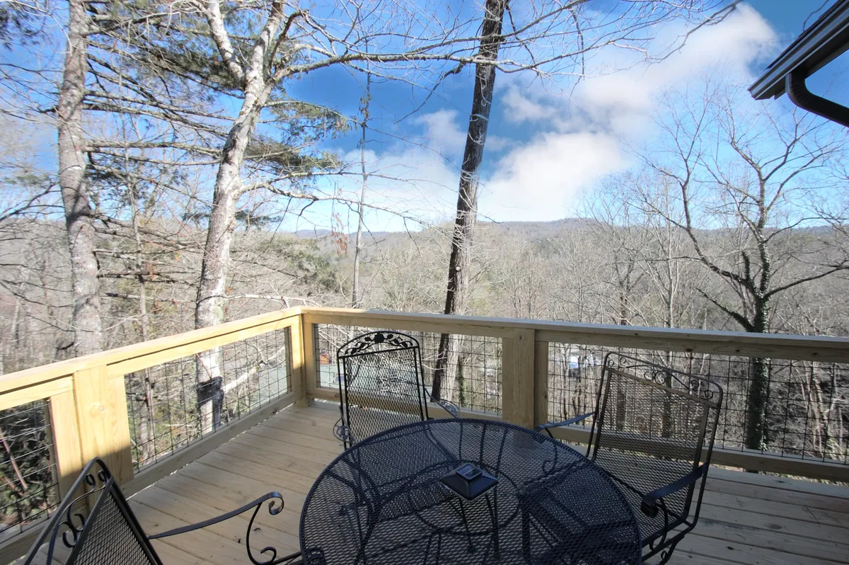 A wooden deck with a metal table and chairs, perfectly positioned to overlook distant mountains and lush tree canopies. The setup is ideal for enjoying morning coffee, group gatherings, and taking in the changing sky.