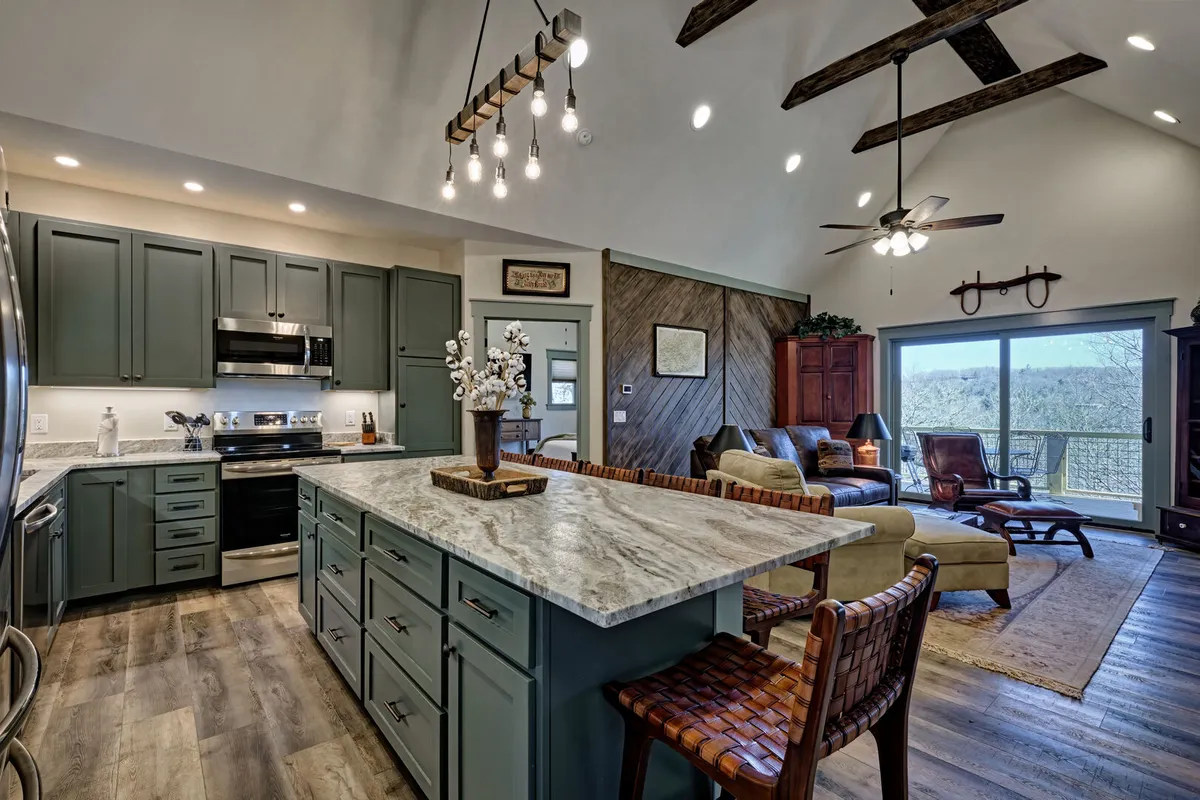 This image showcases a beautifully designed kitchen with a central island featuring bar seating. Large windows flood the space with natural light, highlighting wooden beams overhead and an inviting living area in the background. The seamless visual connection between the kitchen, living area, and outdoors is evident.