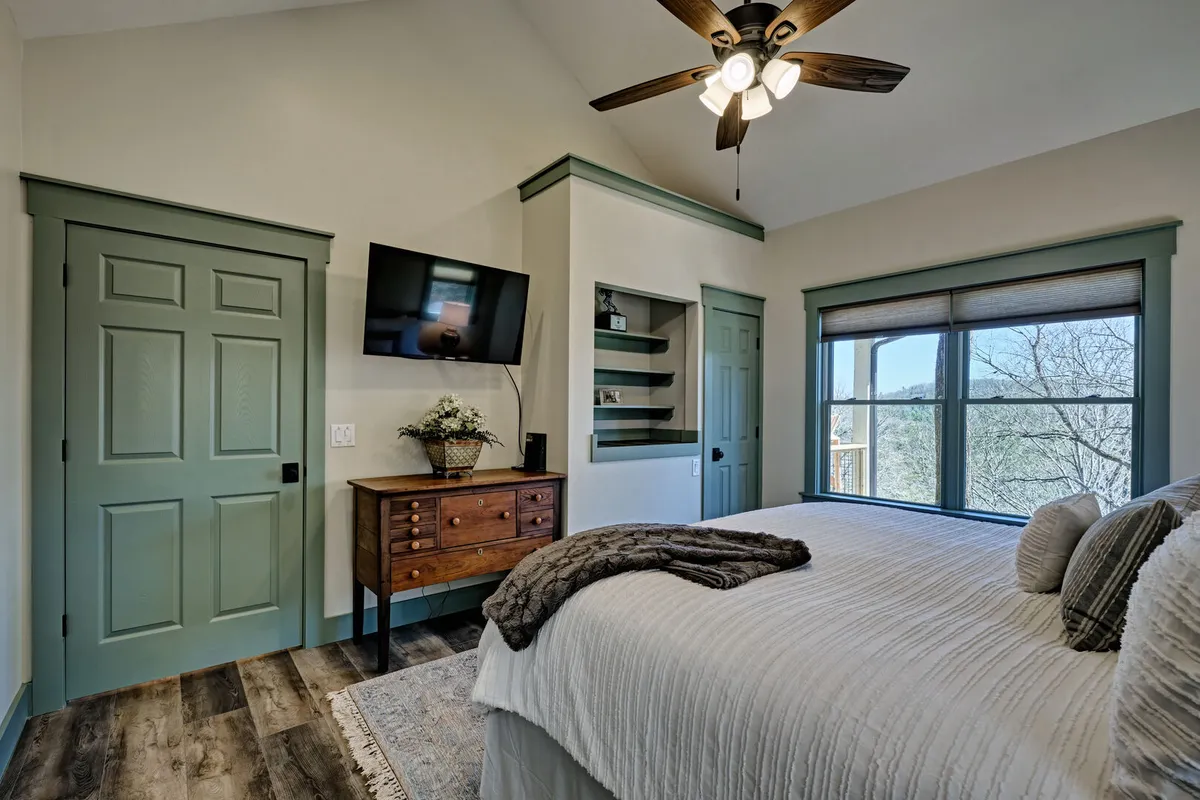 A serene bedroom with a comfortable bed adorned with textured bedding, a large window overlooking a scenic view, a wooden dresser, and a ceiling fan. The peaceful ambiance and soft lighting evoke a sense of restful retreat.