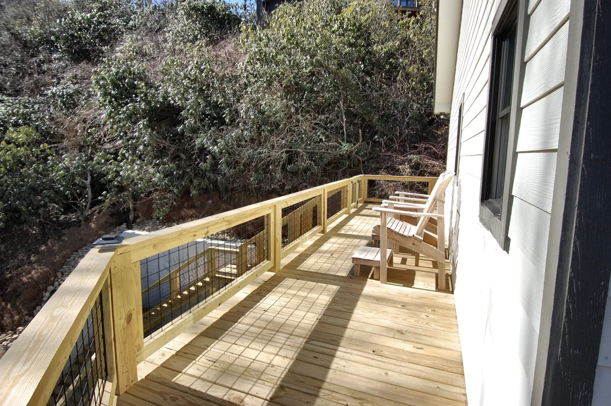 This image features a spacious wooden deck furnished with high Adirondack chairs facing lush greenery. The deck is elevated, framed by secure railings and overlooks the surrounding natural landscape, inviting guests to relax and enjoy the mountain air.