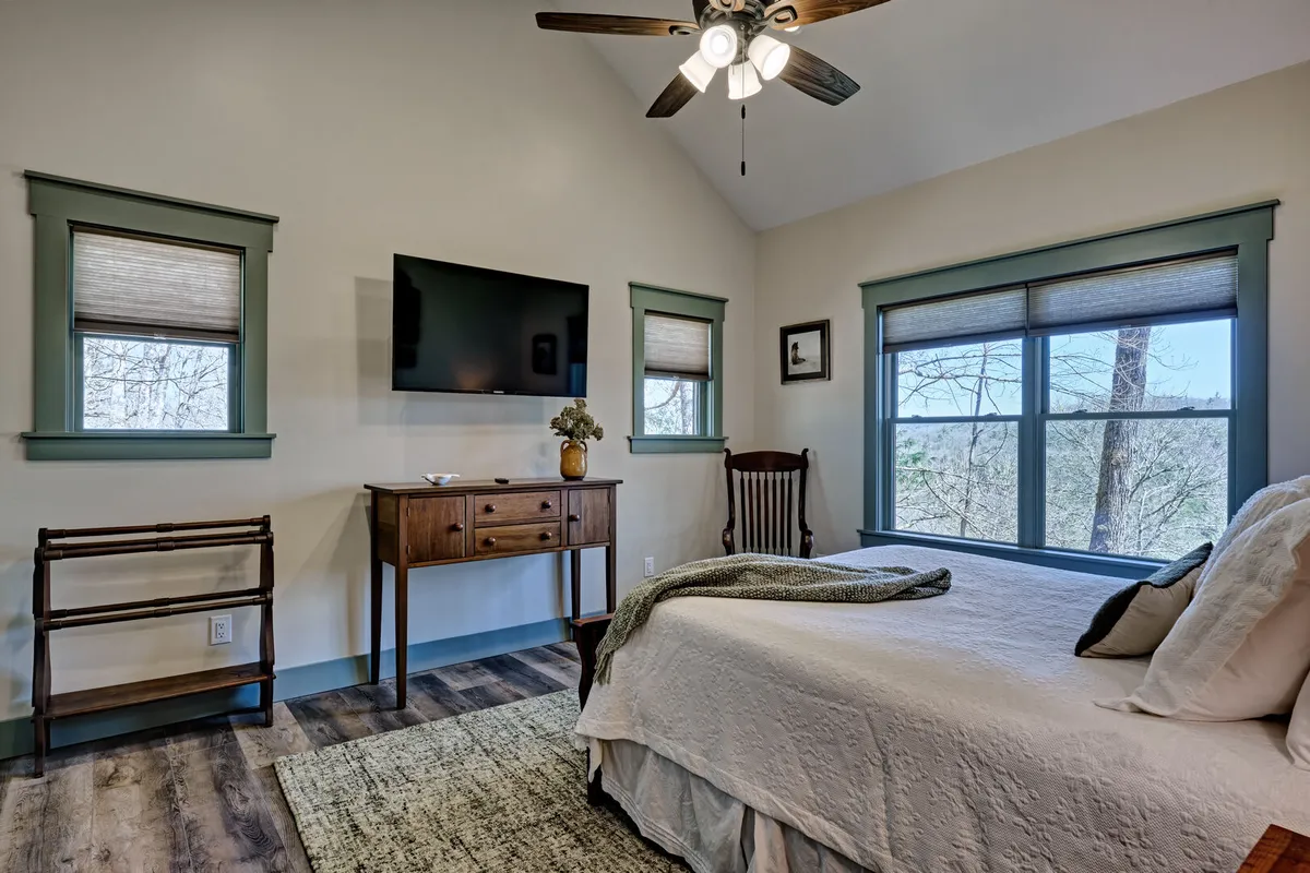 A cozy bedroom featuring a plush bed with a decorative throw, a wooden dresser, a large flat-screen TV mounted on the wall, and expansive windows with views to the outdoors. Natural light fills the space, and the tranquil atmosphere is ideal for relaxing or enjoying a quiet moment.