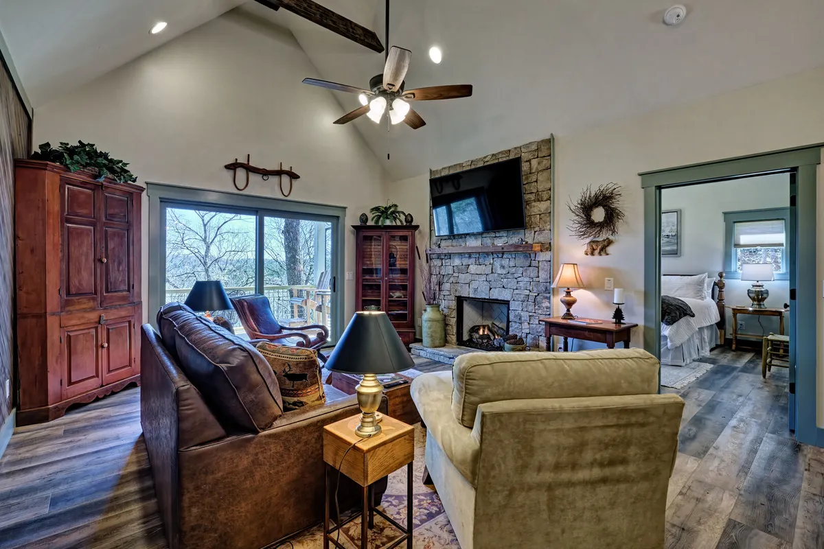 This inviting living area features a prominent stone fireplace framed by large windows that flood the space with natural light. Comfortable seating is arranged for socializing or relaxing, with rustic wooden furniture enhancing the welcoming, communal atmosphere. The overall mood is warm and inviting, perfect for gatherings, movie nights, or cozy evenings by the fire.