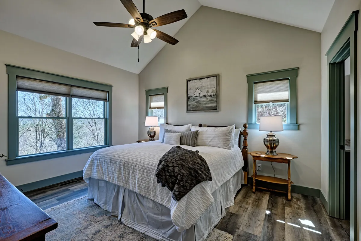 A bright bedroom with large windows letting in natural light, white linens on the bed, and tasteful decorative touches. The expansive windows imply a connection to the outdoors and mountain scenery, aligning with the exploration theme.