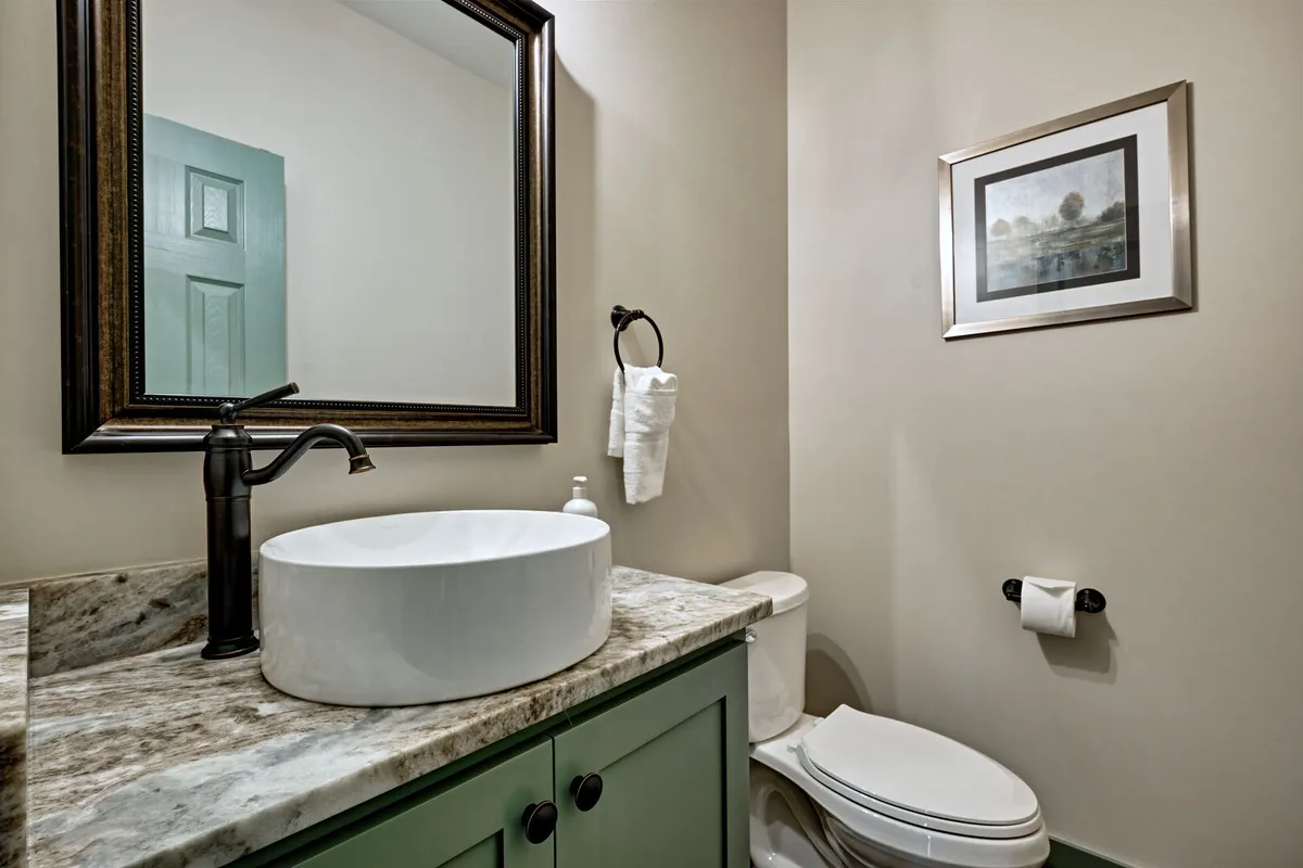 A serene and stylish half bath features an oval sink, decorative mirror, and green cabinetry with framed artwork, offering a peaceful spot for freshening up. The modern yet cozy decor fits seamlessly with the rest of the interior's atmosphere.