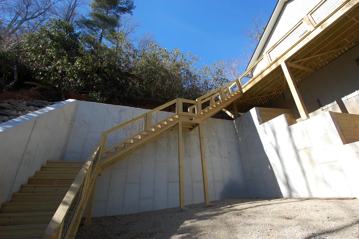 An exterior shot showcasing a wooden staircase leading up to the home, bordered by mature trees and a solid concrete wall. The inviting approach highlights the connection between the house and its natural surroundings.